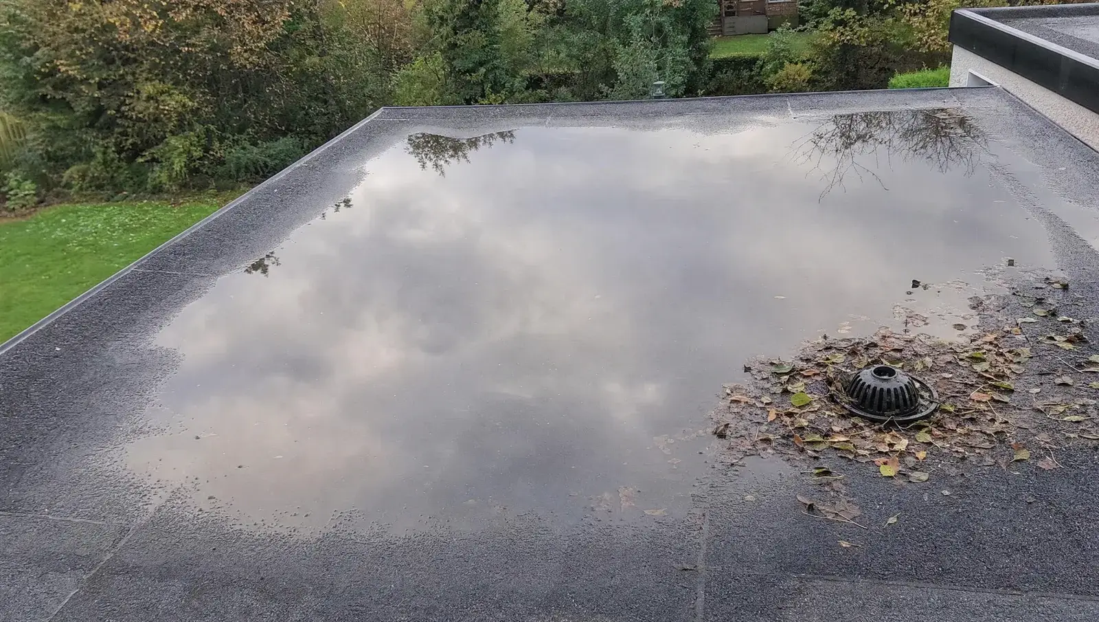 Standing water reflecting the sky on a grey flat roof with leaves gathered around the drain outlet