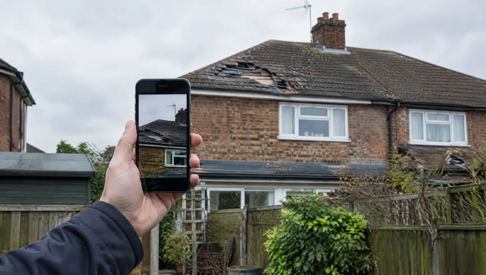 Homeowner photographing storm damage to a roof on a phone from the back garden