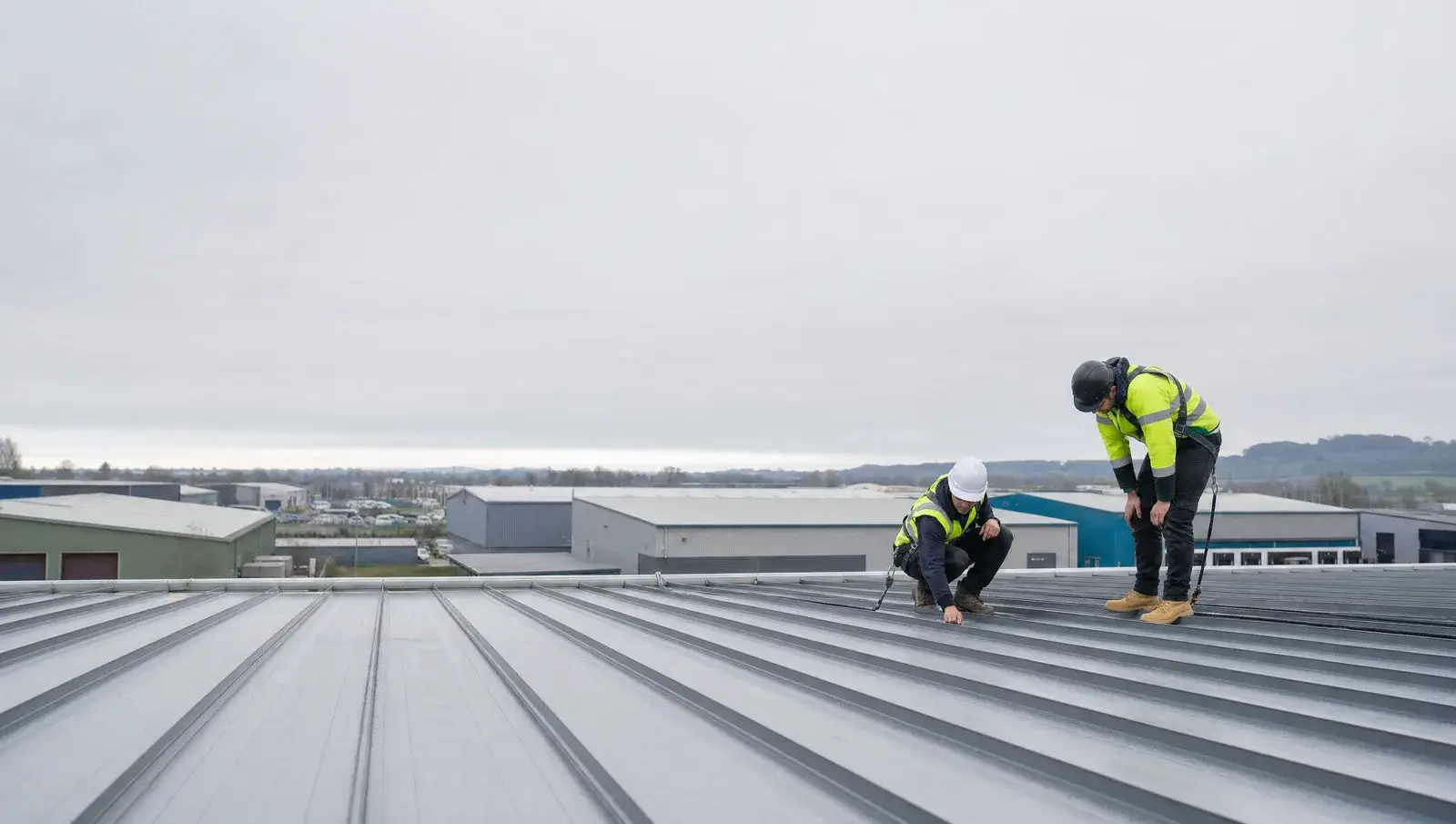 Two roofers in harnesses inspecting standing-seam metal sheets on a large industrial unit roof