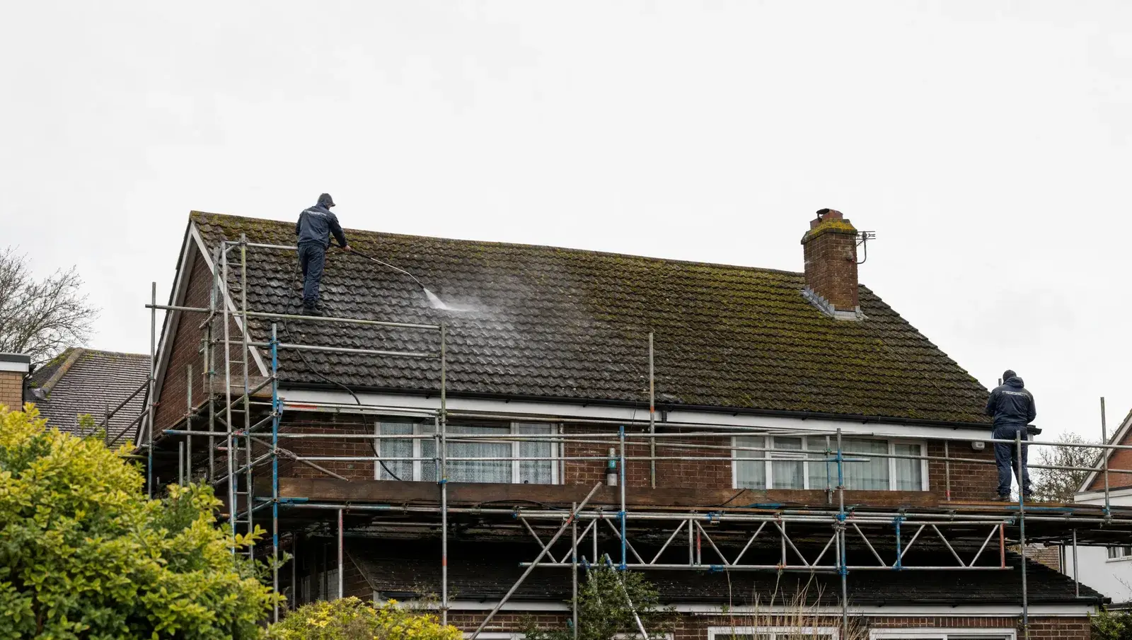 Roof cleaning team soft-washing a large mossy tiled roof from scaffolding